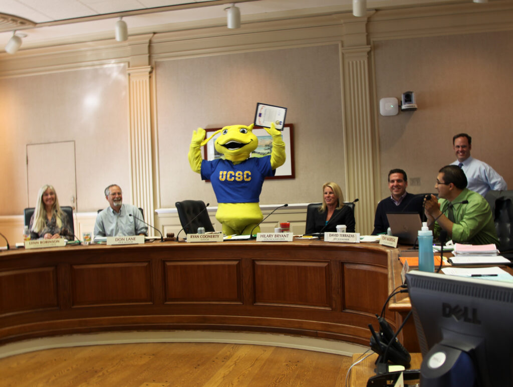 Sammy the mascot in the chambers of the Santa Cruz City council holding their hands overhead in celebration.