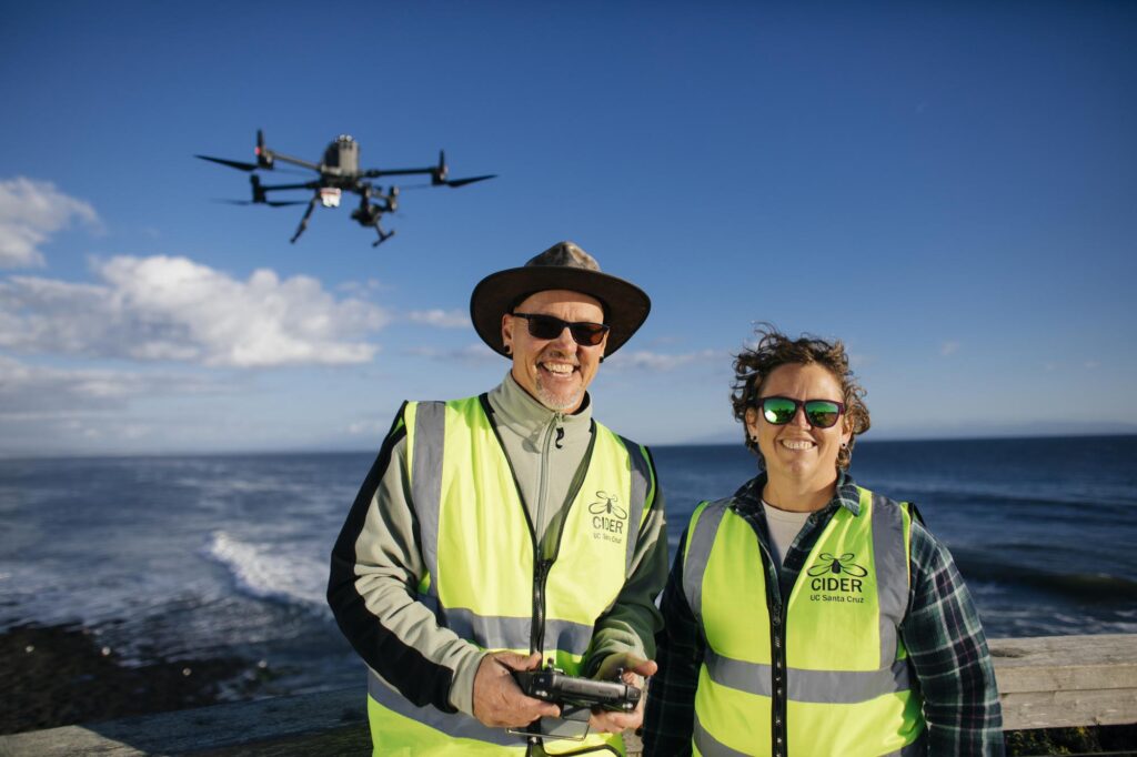 Two people posing for the camera with the ocean in the background and a drone hovering over them.