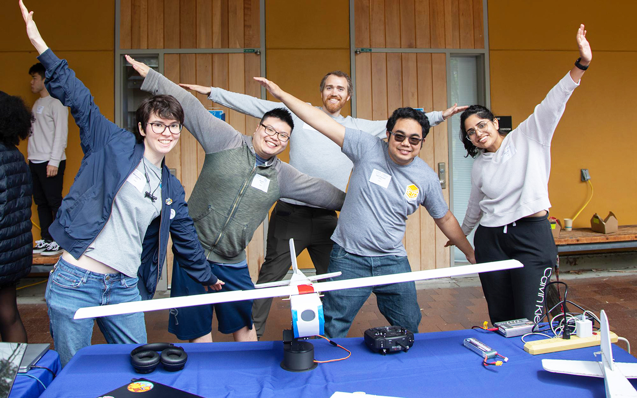 Five students with their arms out like they are flying. They're standing in front of a model plane.