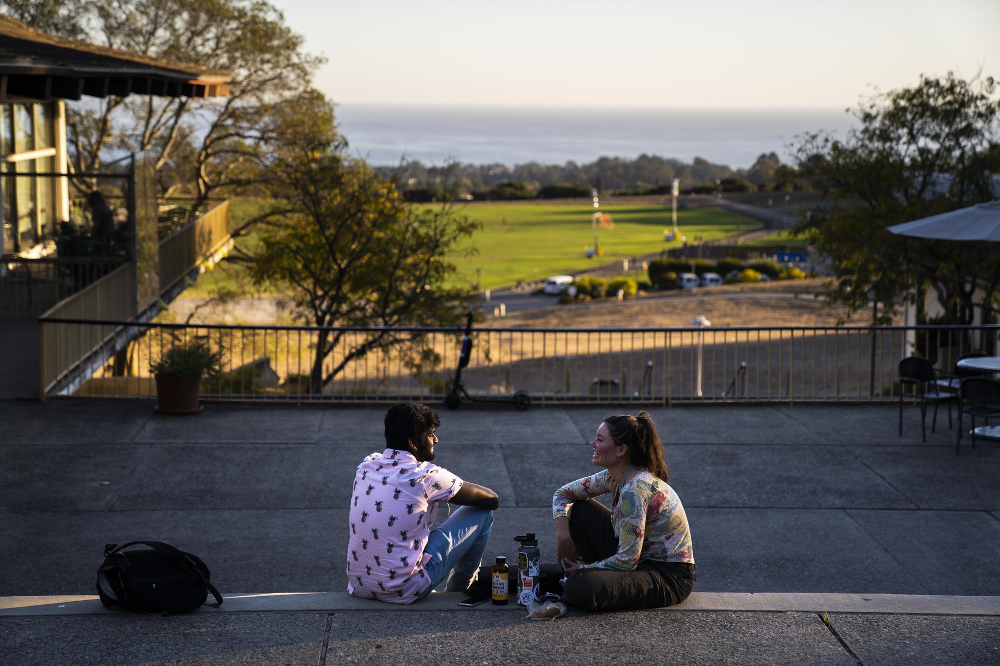 Two people sitting on the steps at Cowell College with a view of the athletics and recreation area in the background.