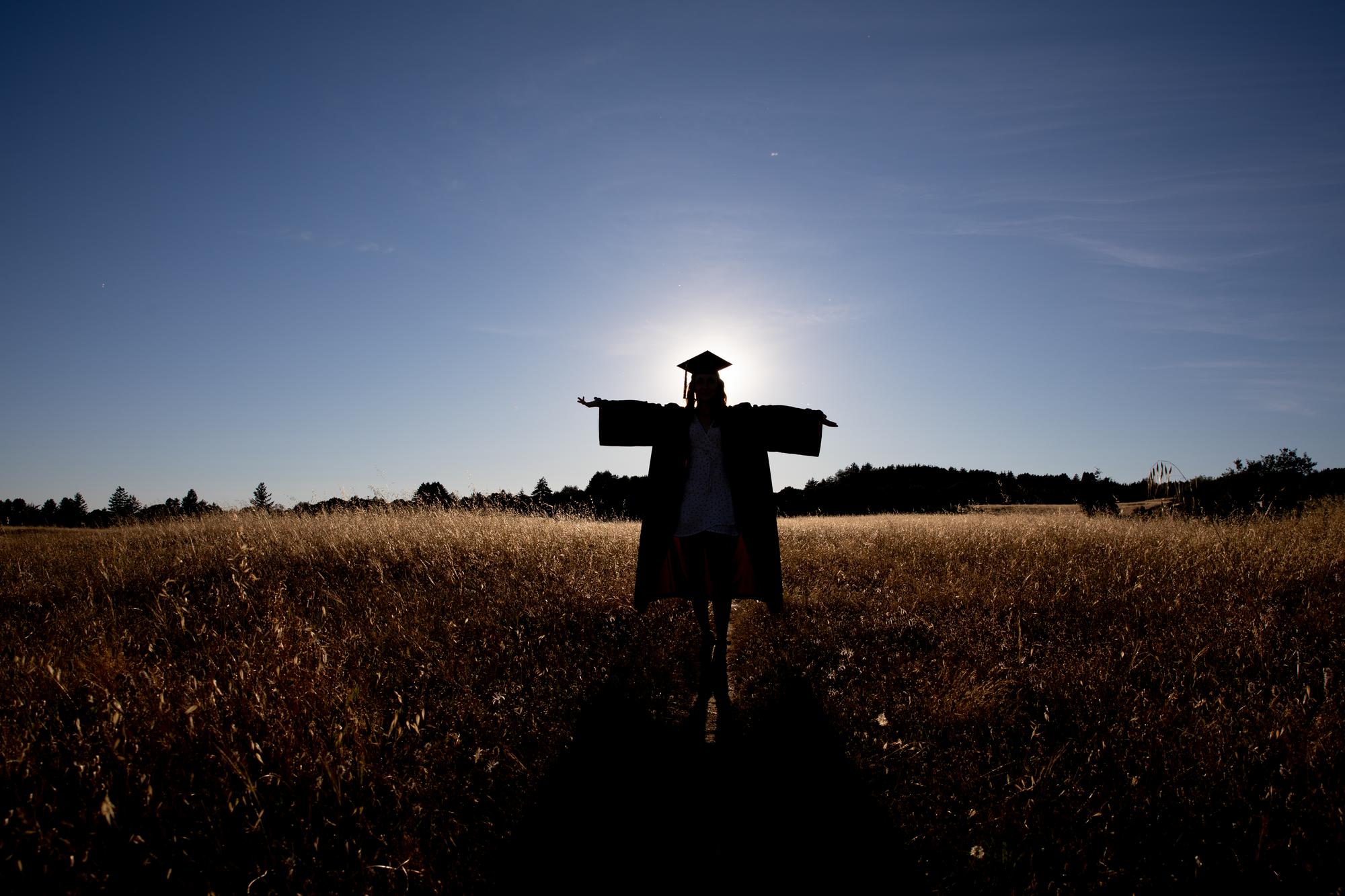Person standing in a field with the sun behind them, wearing their cap and gown.