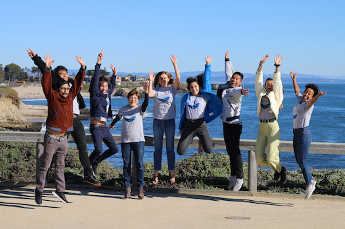 Group of students jumping in the air with the ocean in the background.
