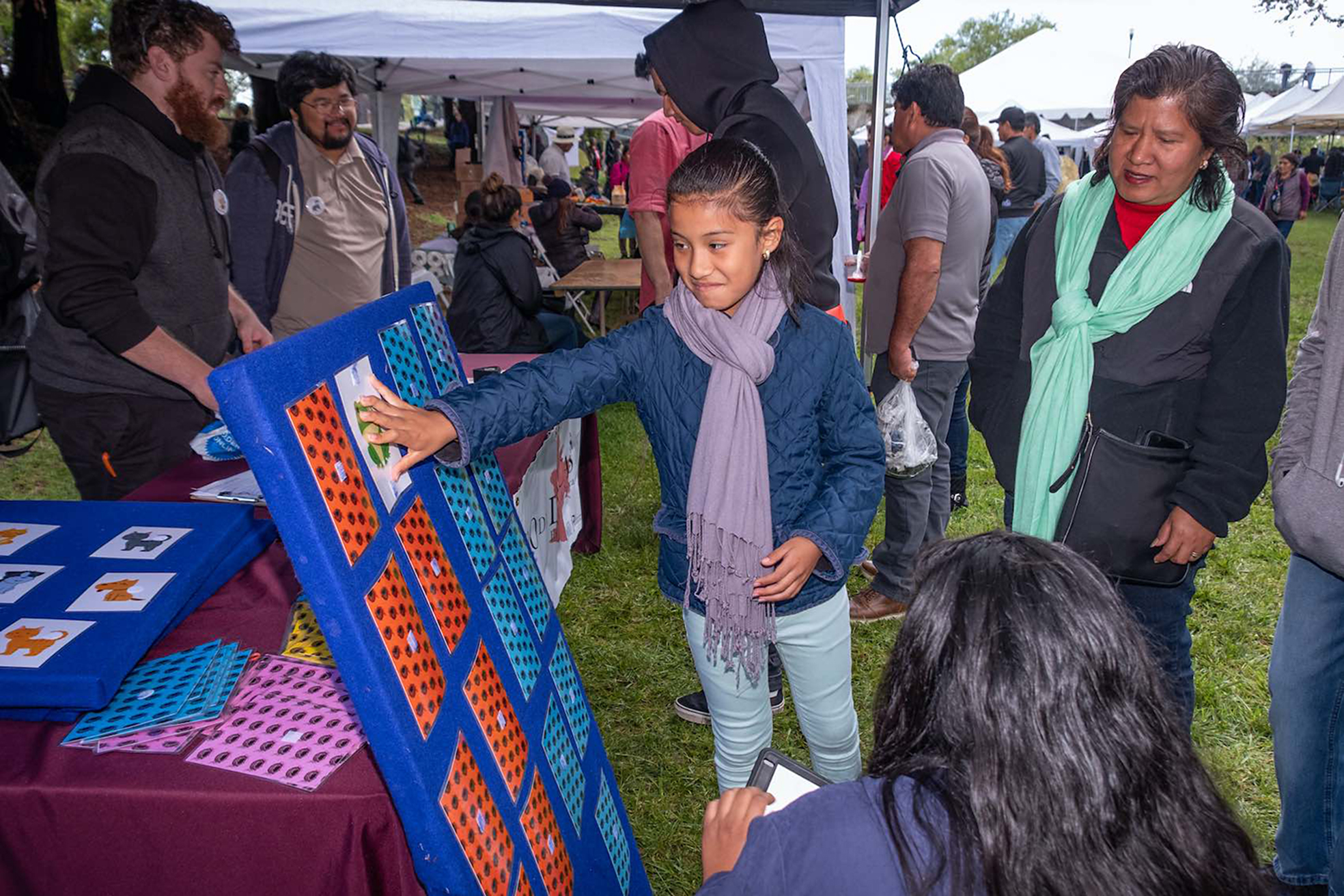 A participant plays a memory matching game at the Nido de Lenguas booth at the Guelaguetza festival organized by Senderos on May 19, 2018 while Linguistics PhD students Max Kaplan (background left) and Andrew Angeles (background right) talk with community members.