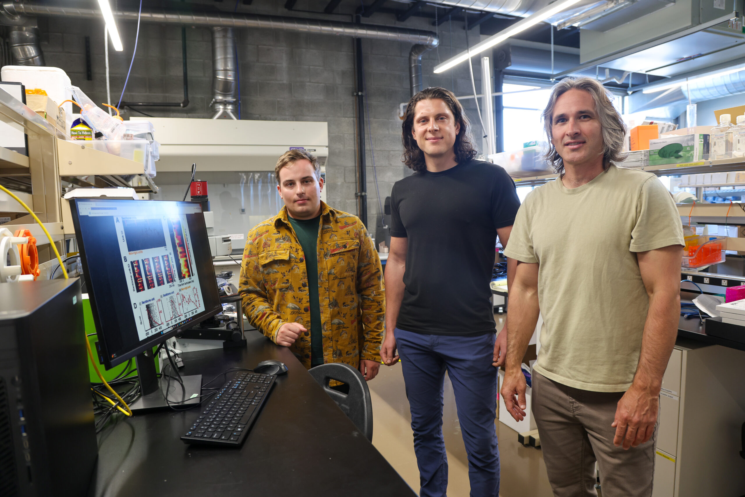 Three researchers pose together in a lab in front of a computer screen showing data visualizations