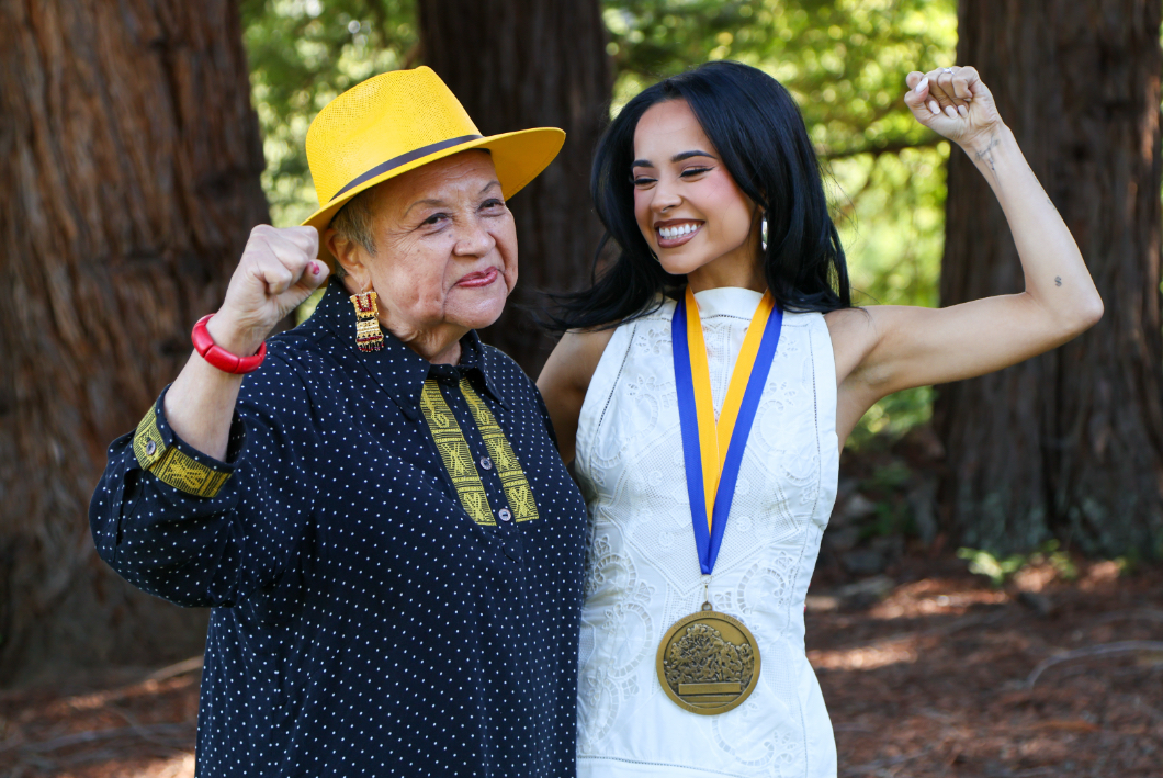 Olga Talamantes in a yellow hat and Becky G with a medal pose outdoors, flexing their arms in celebration. They are standing among tall trees with sunlight filtering through.