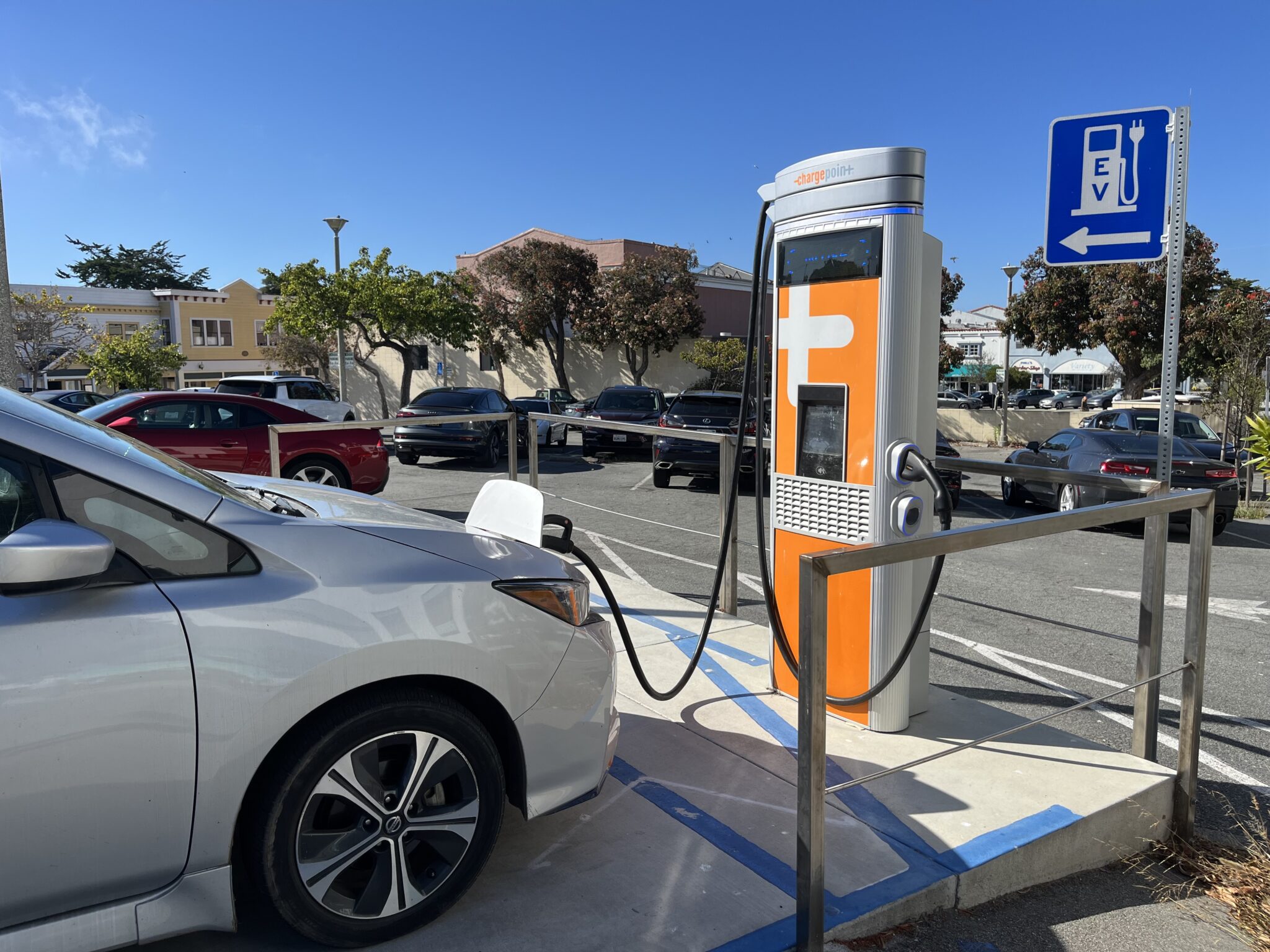 Car plugged into a public faster charger in a parking lot in a downtown area, with an "EV" sign. 