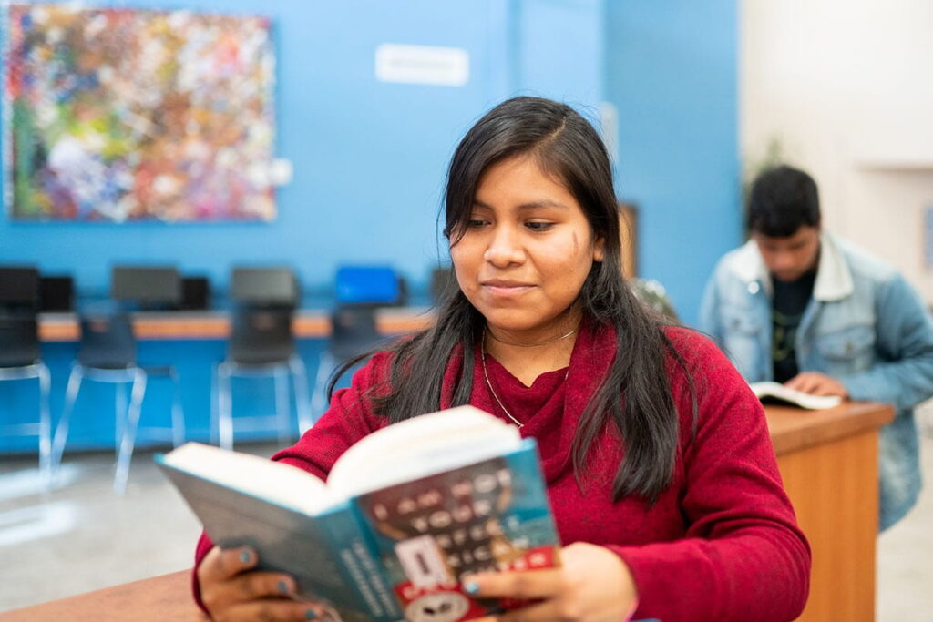 A female student reading an open book in a classroom setting.