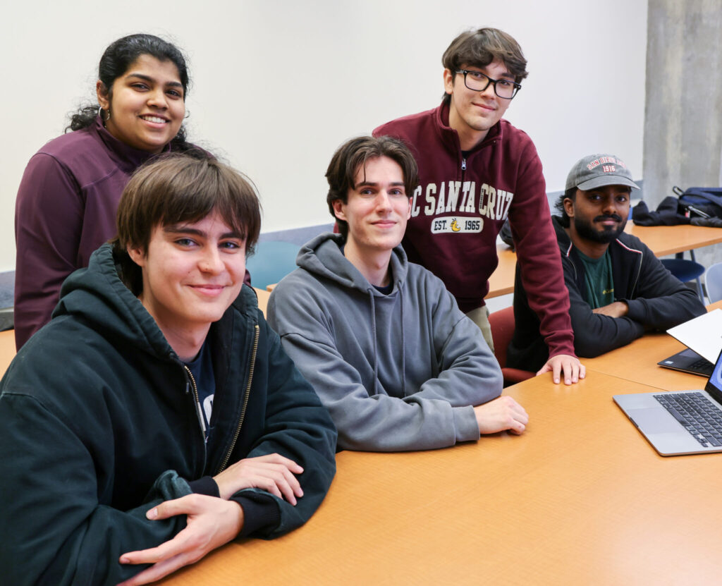 Four students sitting together around a table with laptops in a classroom.