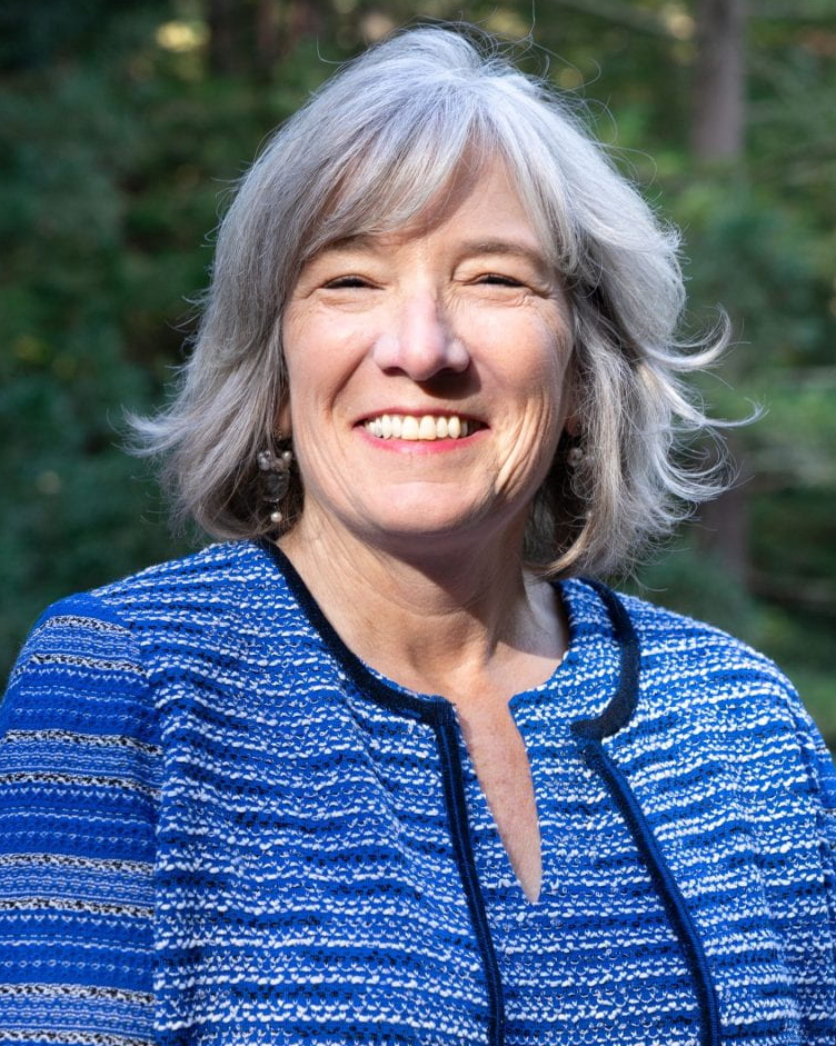 A smiling woman with gray hair wears a blue patterned jacket and stands outdoors with trees in the background.