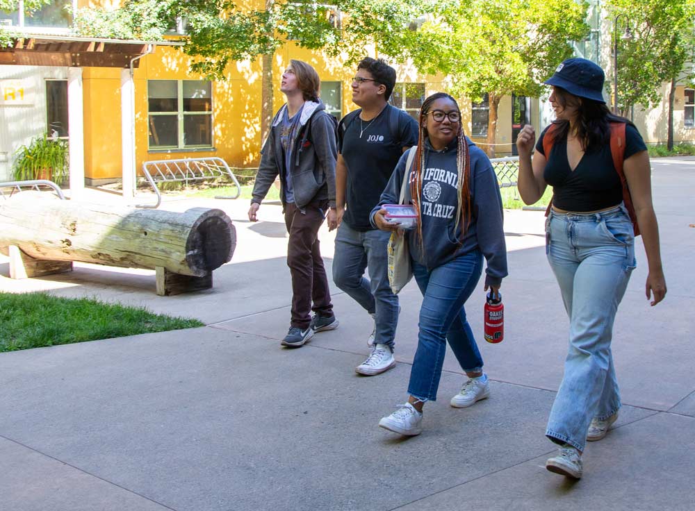 A diverse group of four students walking and talking outdoors on a sunlit campus plaza in front of yellow campus buildings.