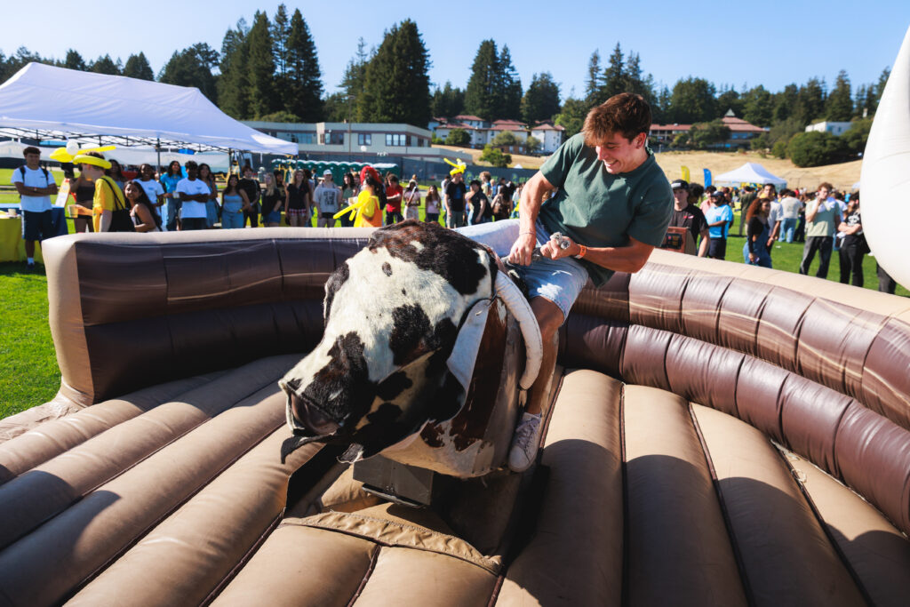 A student riding a mechanical bull at an outdoor campus event.